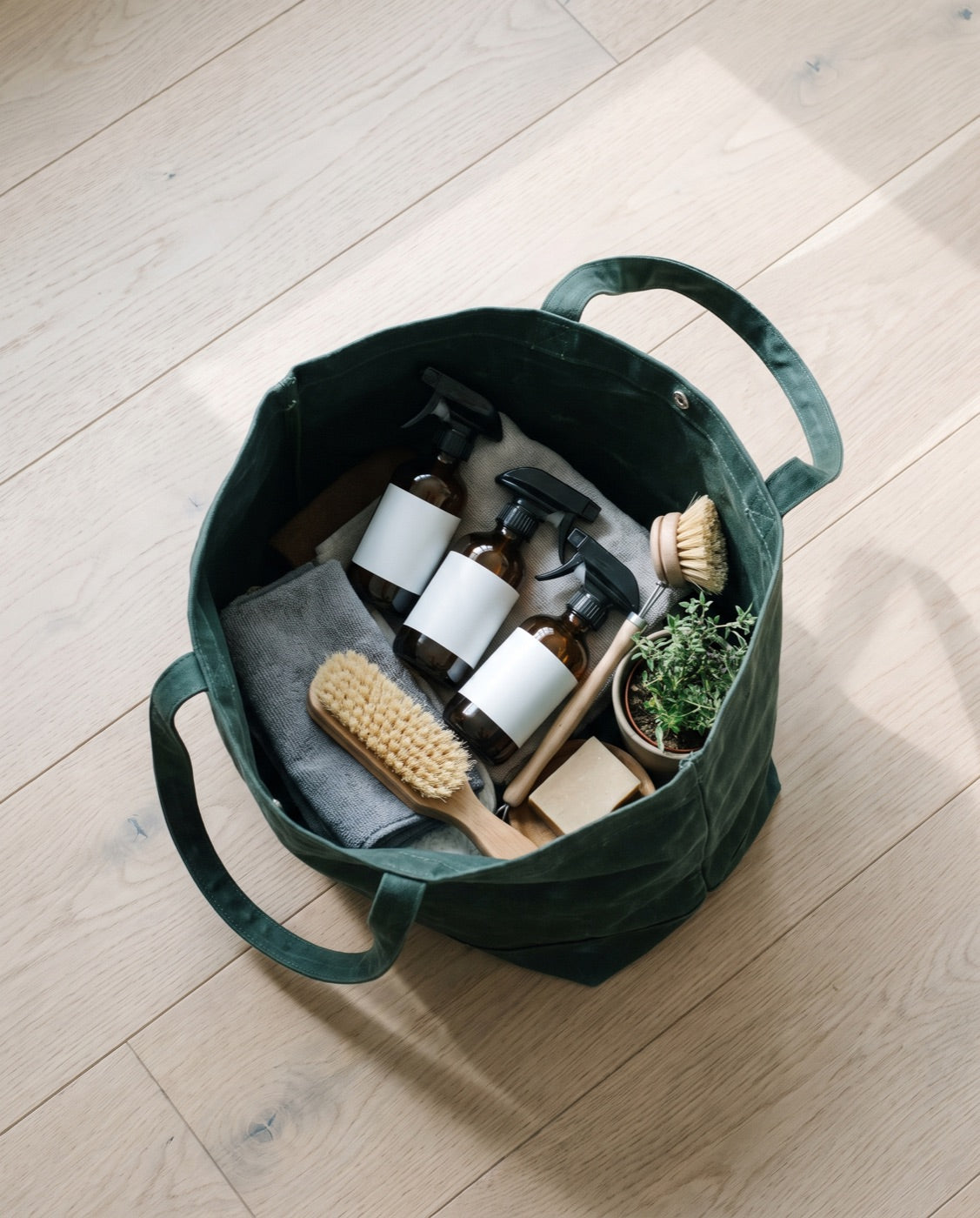 A green canvas tote of cleaning supplies on white oak floorboards