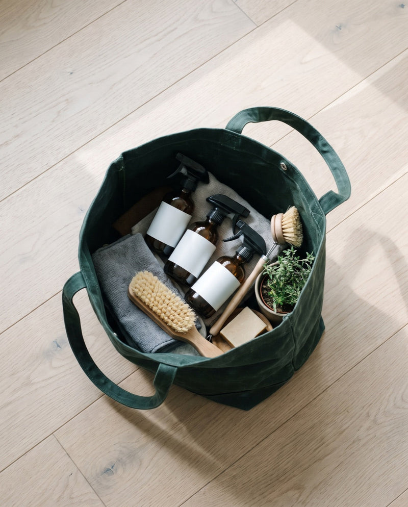A green canvas tote of cleaning supplies on white oak floorboards